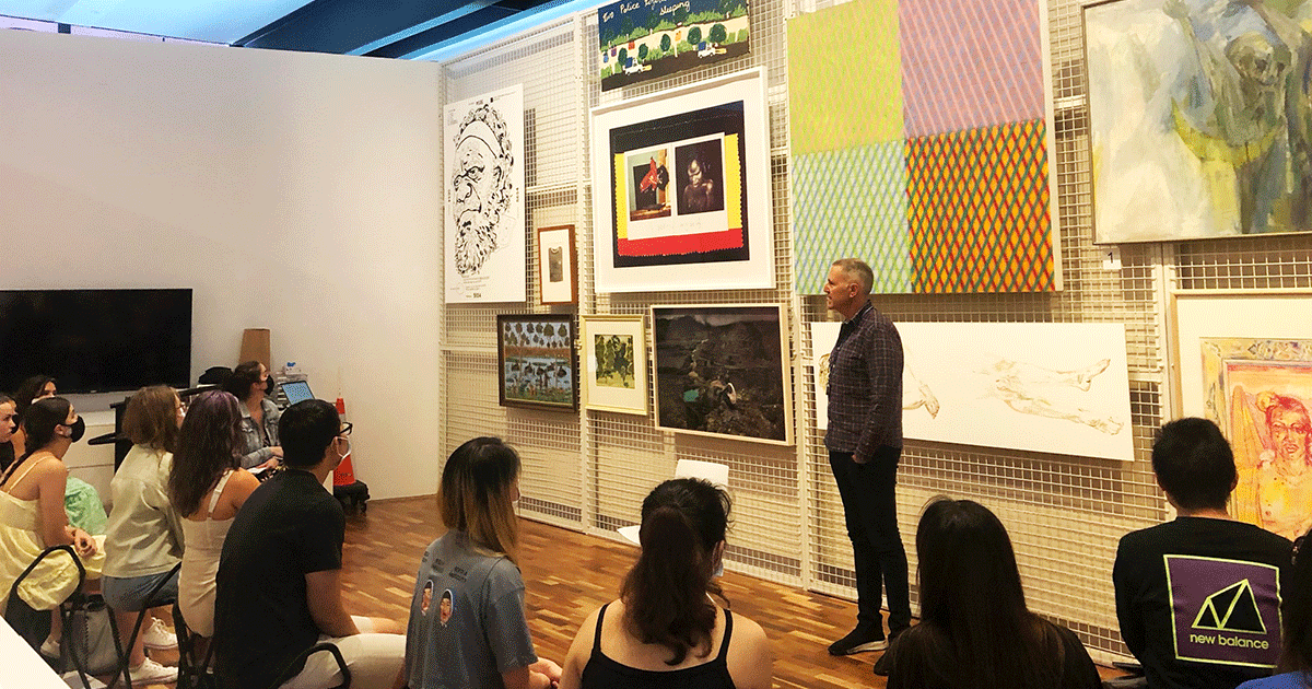 A person giving a talk to a group of students in front of a wall filled with artworks